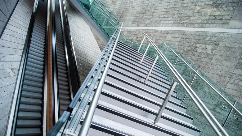 Das Foto zeigt zwei Rolltreppen sowie eine Treppe, die über die beiden Rolltreppen führt. Die Rolltreppen laufen gerade aus.  Die moderne aus Metall bestehende Treppe überquert die Rolltreppen, jedoch versetzt im Zickzack. 